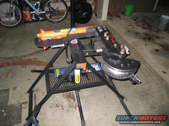 Orange hydraulic pipe bender on a black metal stand, with a metal tube in the bending die