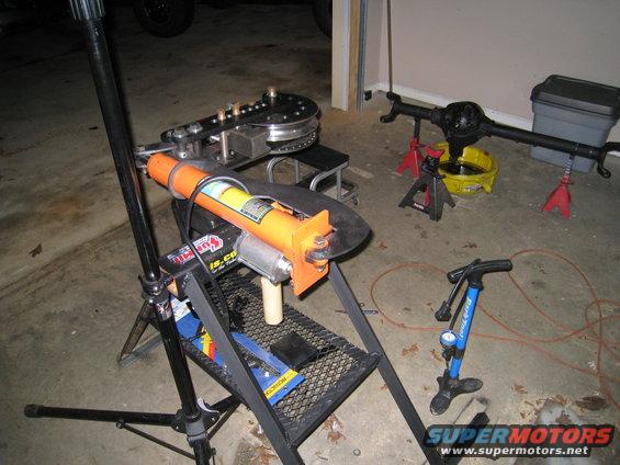Pipe bender with orange hydraulic ram and a rear axle assembly on jack stands