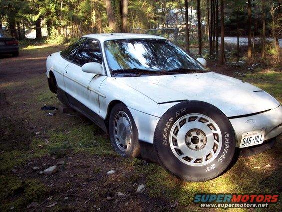 White 1991 Ford Probe with a spare tire in front, parked in a wooded area. Another dark car is visible in the background