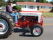 1966 Ford Ranchero - A man in overalls and a hat drives a red and white Ford 801 PowerMaster tractor down a street.