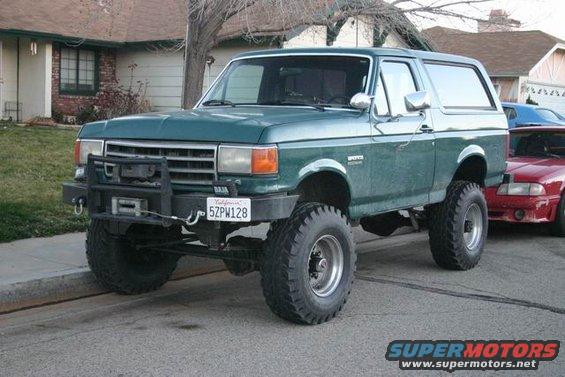 1991 Ford Bronco, lifted with large tires, aftermarket front bumper, winch, and California license plate 5ZPW128. Red car visible behind it