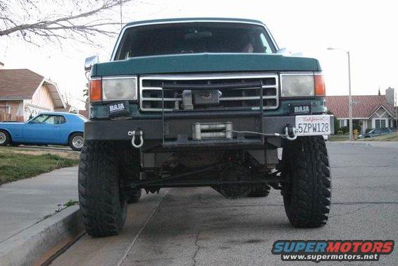 Green lifted pickup truck with winch and Baja stickers, parked on street next to a blue car