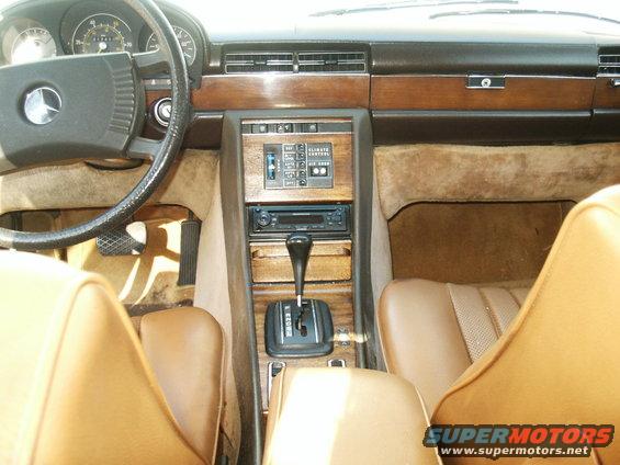 Car interior with wood trim, tan seats, and a Mercedes-Benz steering wheel. The center console shows climate controls and a gear shifter