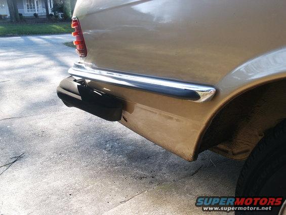 Rear bumper and taillight of a tan car. Chrome trim above bumper