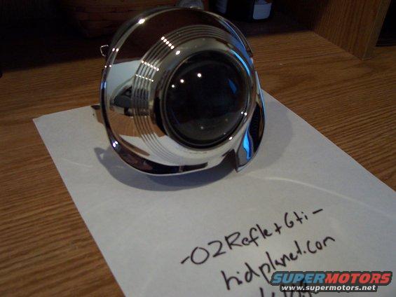 Chrome headlight projector with black lens on white paper, text reads "-02Refle+6ti- hidplanet.com"