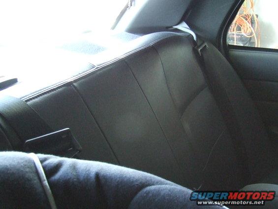 Back seat of a car with black leather upholstery and a seatbelt. A person's shoulder is visible in the foreground