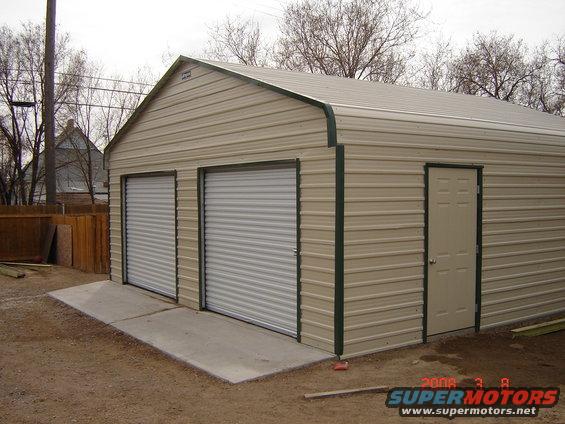 Tan metal garage with green trim, two white roll-up doors, and a beige side door