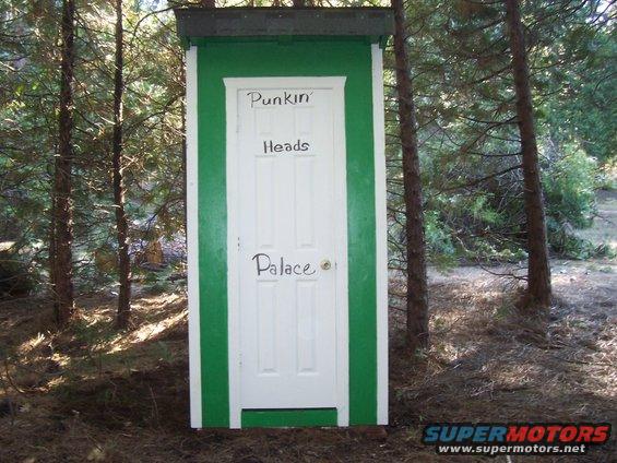 Green and white outhouse with 'Punkin Heads Palace' written on the door, set in a wooded area