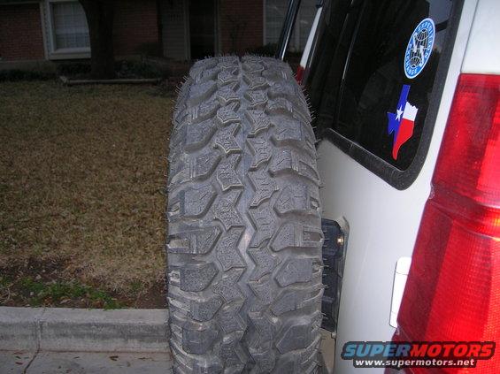 Spare tire with aggressive tread mounted on the rear of a white SUV, with Texas flag sticker on window