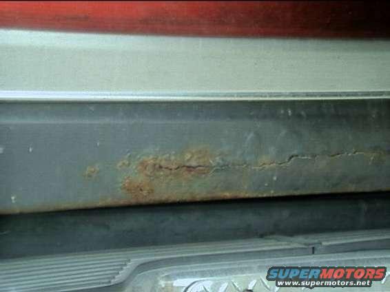 Close-up of a cracked and rusted metal panel on a vehicle, with a red stripe above and a ribbed section below