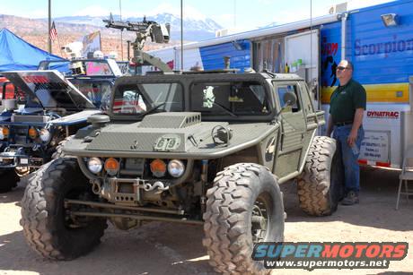 Olive green military vehicle with machine gun, large tires, and a man standing nearby