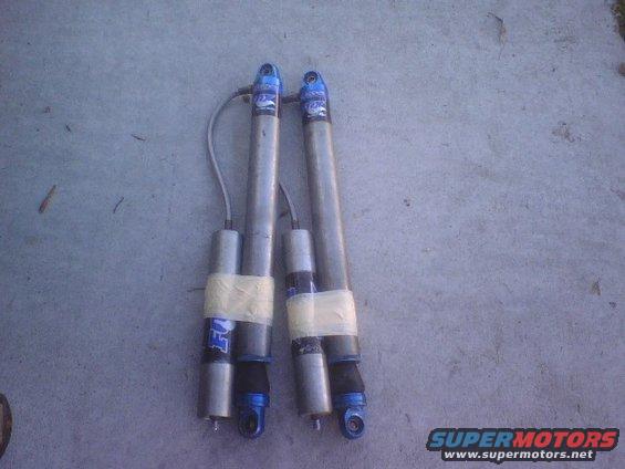 Two Fox shock absorbers with blue tops and silver bodies, taped with white tape