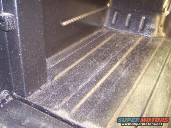 Close-up of a truck bed floor with ribbed metal and some dust. A black vertical panel is on the left
