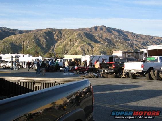 Swap meet scene with trucks, trailers, and people gathered near mountains