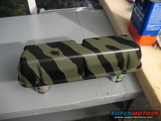 Camshaft cover painted with olive drab and black tiger stripes, resting on cans