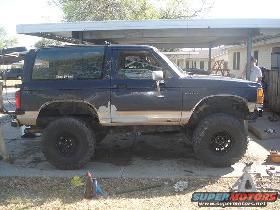 Side view of a dark blue Ford Bronco II with large off-road tires and some body damage. It has a tan stripe and a sticker on the rear window