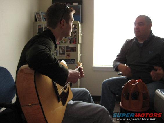 guy playing acoustic guitar while another man watches in a room with a window