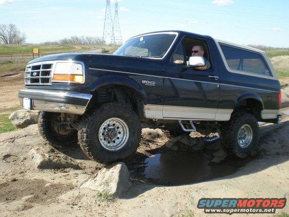 Dark blue and tan Ford Bronco driving over rocks and through mud