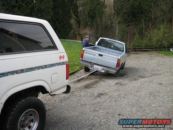 White Ford Bronco towing a silver Nissan pickup truck on a gravel driveway