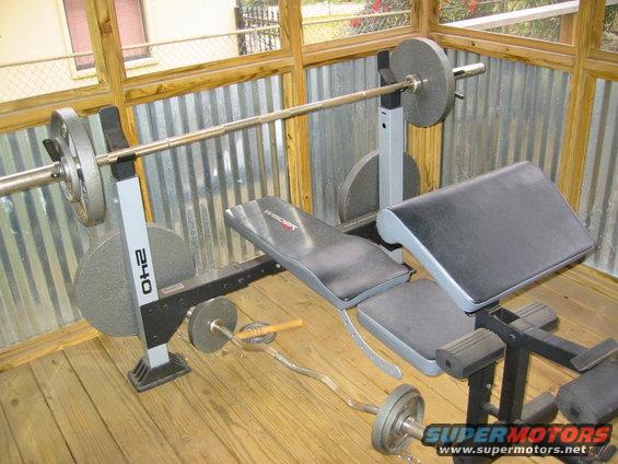 Weight bench with barbells and weight plates on wooden floor, corrugated metal wall behind