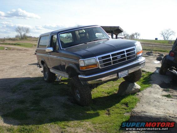 Dark blue Ford Bronco with lifted suspension, front tire on a rock. Another SUV is visible to the right