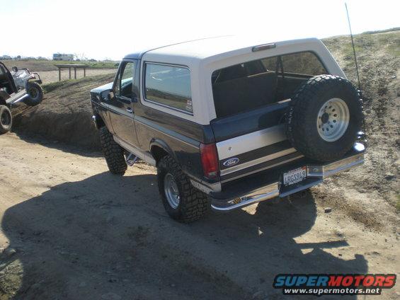 Rear view of a 1993 Ford Bronco with a spare tire mounted on the back, driving on a dirt road. Another ATV is visible on a hill