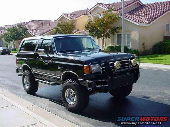 Black 1987 Ford Bronco with custom grille guard and smiley face lights, parked on a suburban street