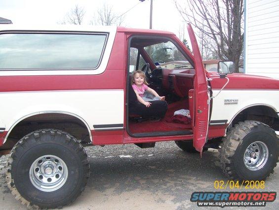 1988 Ford Bronco, red and white, with a child sitting in the driver's seat and the passenger door open