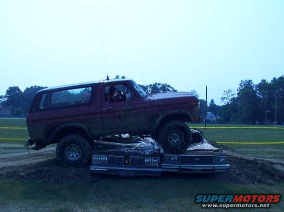 Red Ford Bronco driving over two sedans in a muddy field