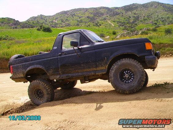 Black Ford Bronco with lifted suspension driving on a dirt road with hills in the background. The front passenger side tire is turned
