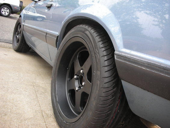 Car side view showing black five-spoke rims and tires. Another car is parked behind it