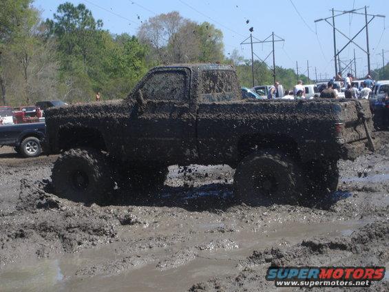 Mud-covered pickup truck driving through a mud pit with other vehicles and spectators in the background