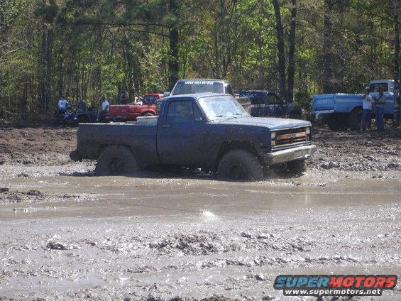 Blue pickup truck with large tires stuck deep in mud, other vehicles and people in background