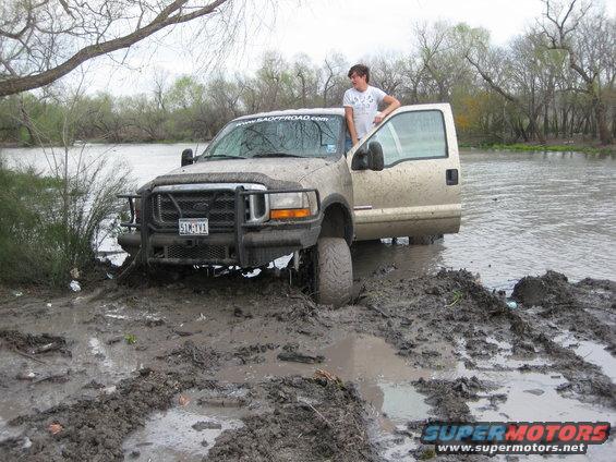 Mud-covered tan Ford F-250 Super Duty with open driver door stuck in mud near water