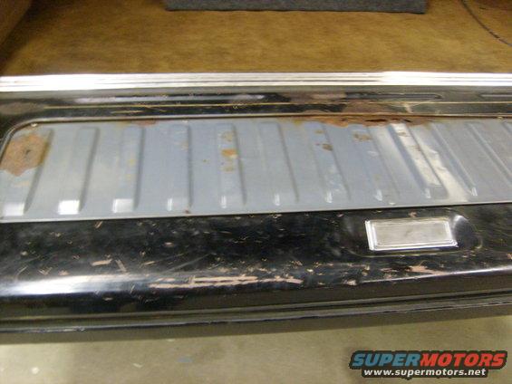 Close-up of a vehicle's rear cargo area, showing a gray ribbed panel with rust spots and a black painted surface with scratches. There's a chrome trim piece on the right