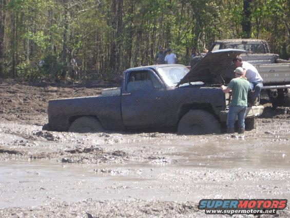Mud-covered dark blue pickup truck with hood open, stuck in mud. Two people examine engine. Another truck visible behind