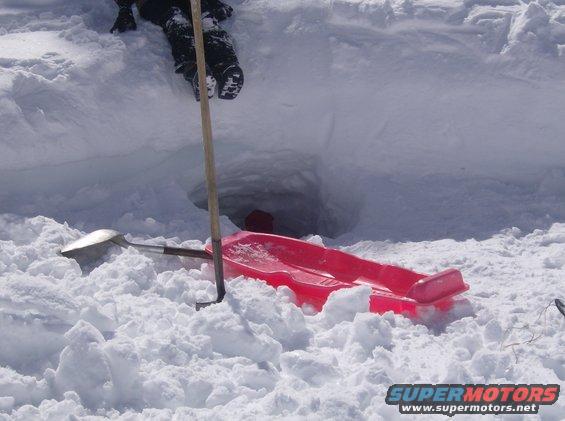 Red plastic sled and shovels in deep snow next to a dug-out hole