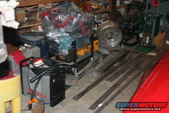 Engine covered in plastic wrap, a metal wheel, welding machine, and metal bars are in a garage