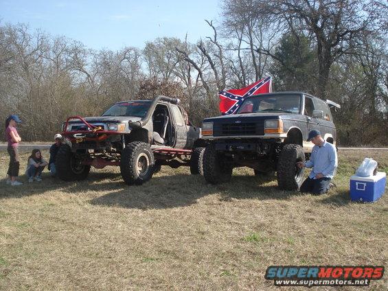 1989 Ford Bronco II and a lifted pickup truck parked on grass, with people nearby and a Confederate flag on the Bronco II