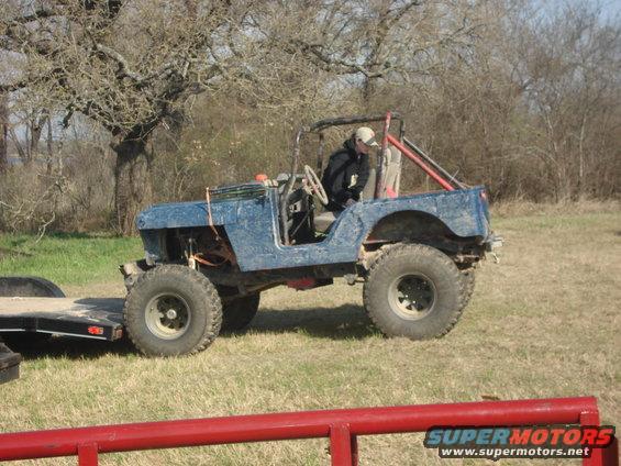 Blue off-road Jeep with roll cage, large tires, and a trailer attached. Person is in the driver's seat