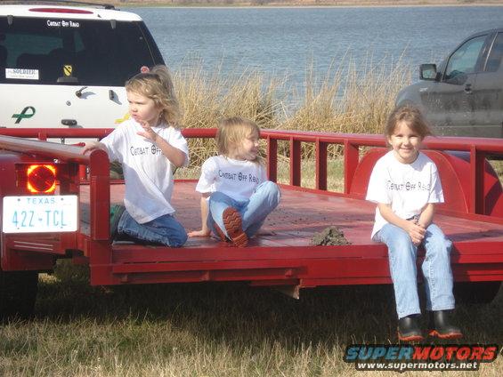 Three young girls sit on a red trailer. A white SUV with stickers is behind them, and a dark SUV is to the right. Water and dry grass are in the background