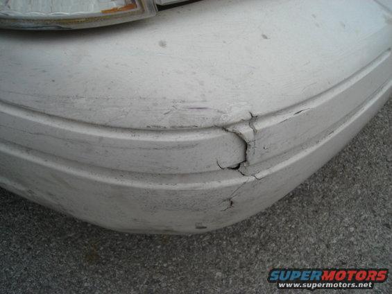 White car bumper with significant cracks and scuffs, showing a portion of a headlight