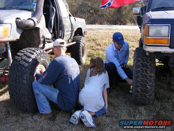 Two men and a girl crouch near large tires of modified off-road trucks; one man checks tire pressure