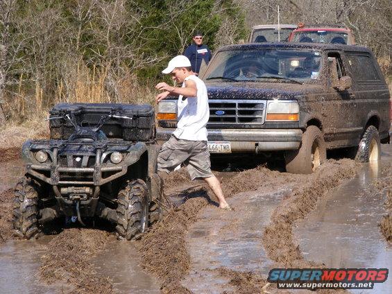 Mud-covered 1992 Ford Bronco stuck in mud, with an ATV next to it. A person is stepping out of the Bronco