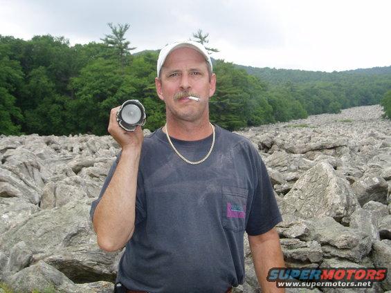 Man in a dark t-shirt and white cap holds a flashlight, smoking a cigarette, with a rocky landscape behind him