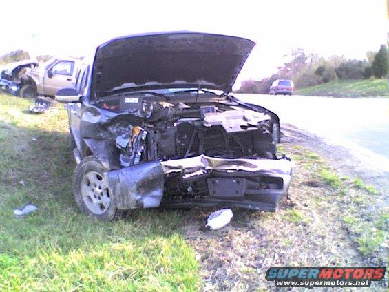 Wrecked dark pickup truck with hood open, front end damaged. Another wrecked truck is visible in the background