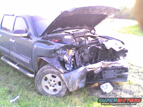 Front end of a severely damaged pickup truck with the hood open, showing crumpled metal and engine components