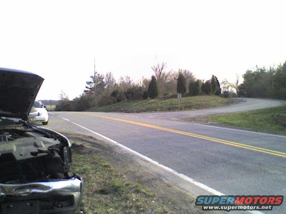 Dark vehicle front with hood open, white car in background, roadside
