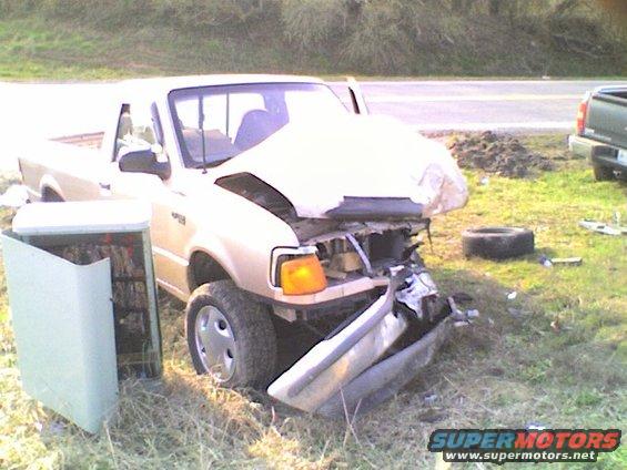 Gold pickup truck with a severely damaged front end, hood crumpled, bumper hanging. Open door reveals contents. A spare tire lies nearby