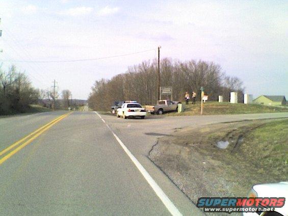 Police car and pickup truck parked on the side of a rural road with a few people standing nearby